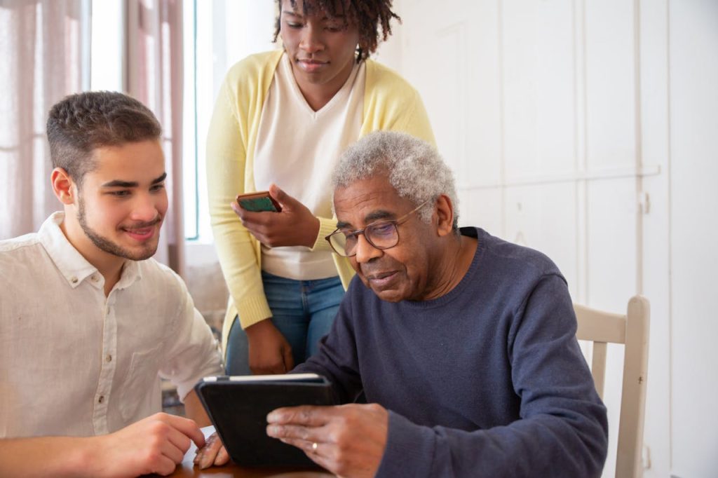 An Elderly Man in Blue Sweater Holding a Tablet while Talking to the People Beside Him